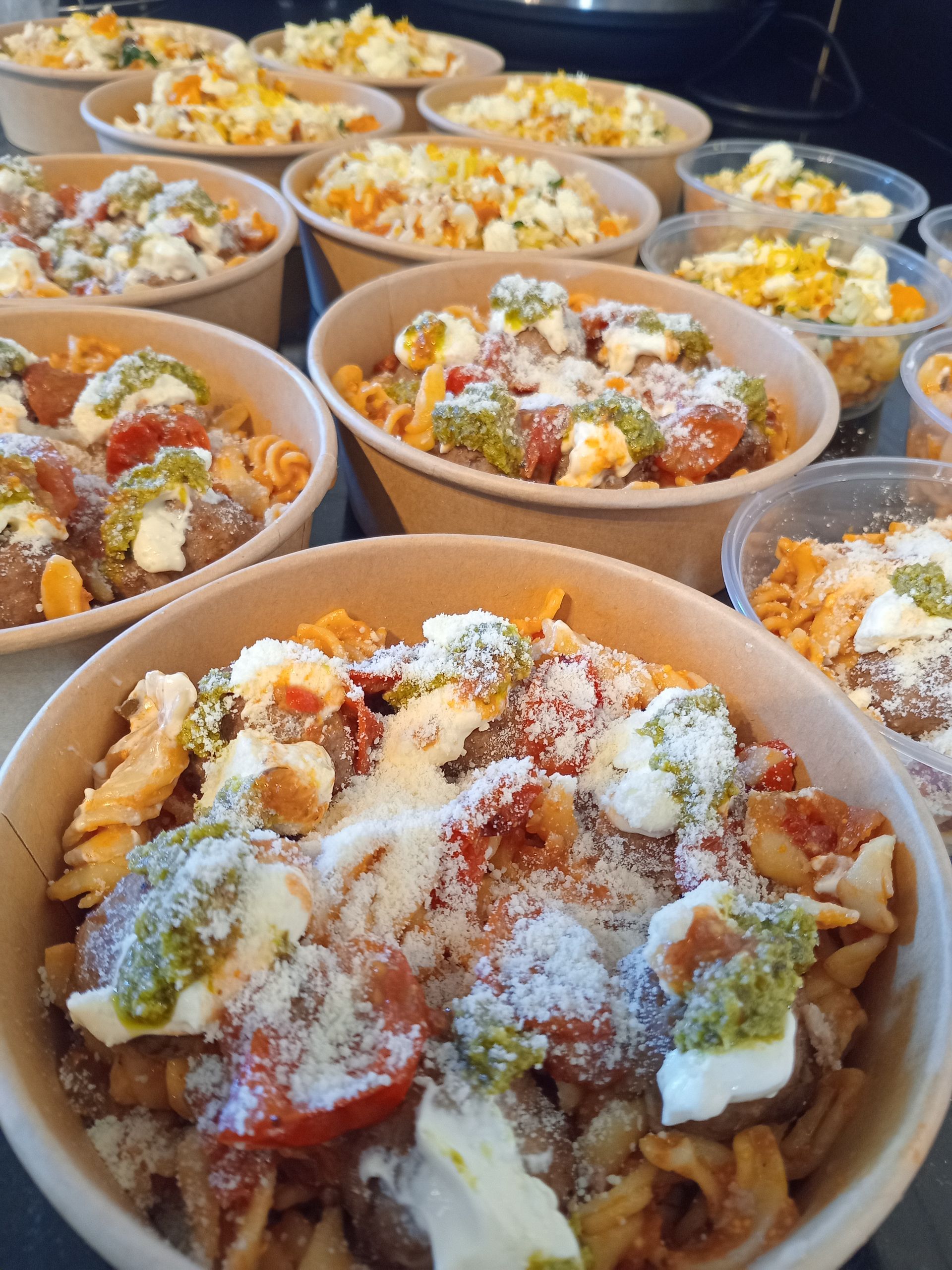 Pasta dishes in paper bowls, various toppings like pesto, cheese, tomatoes.