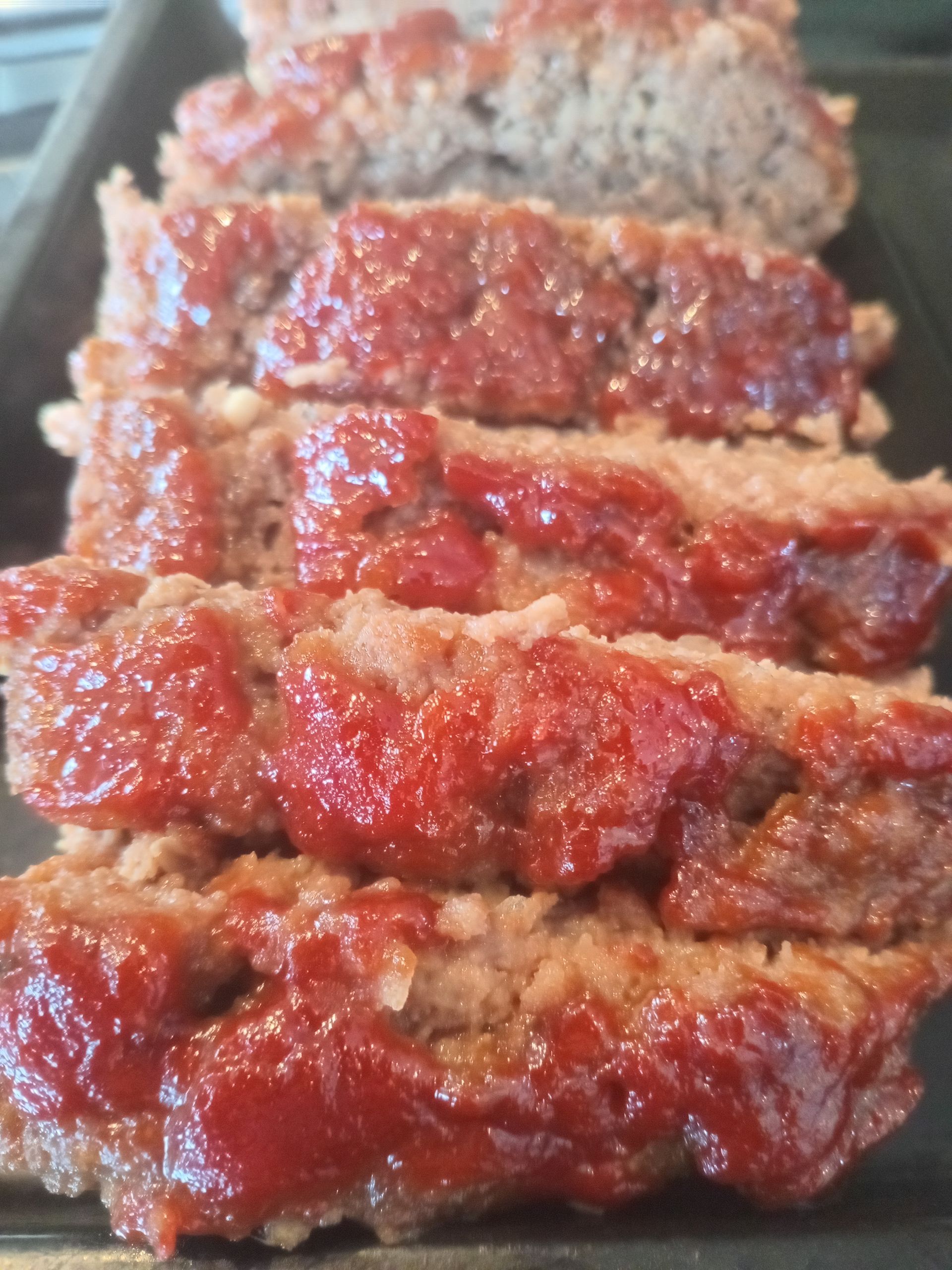 Slices of meatloaf, topped with a red sauce, on a black baking sheet.