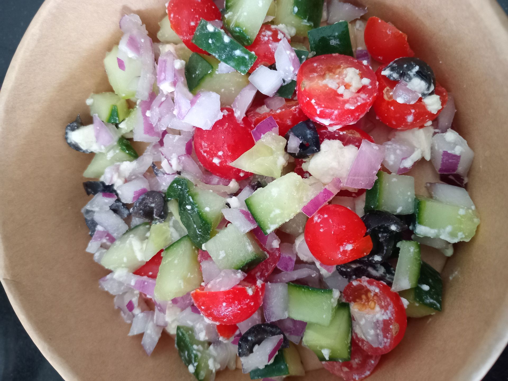 Chopped cucumber, cherry tomatoes, red onion, and black olives in a brown bowl.