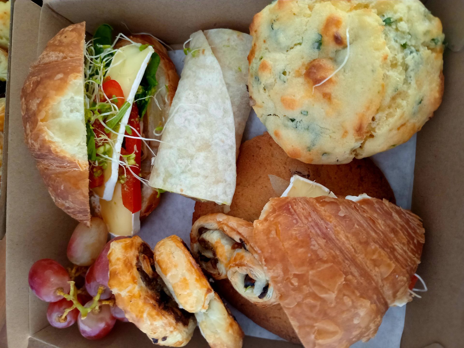 Boxed lunch with croissant, wrap, muffin, grapes, and pastry.