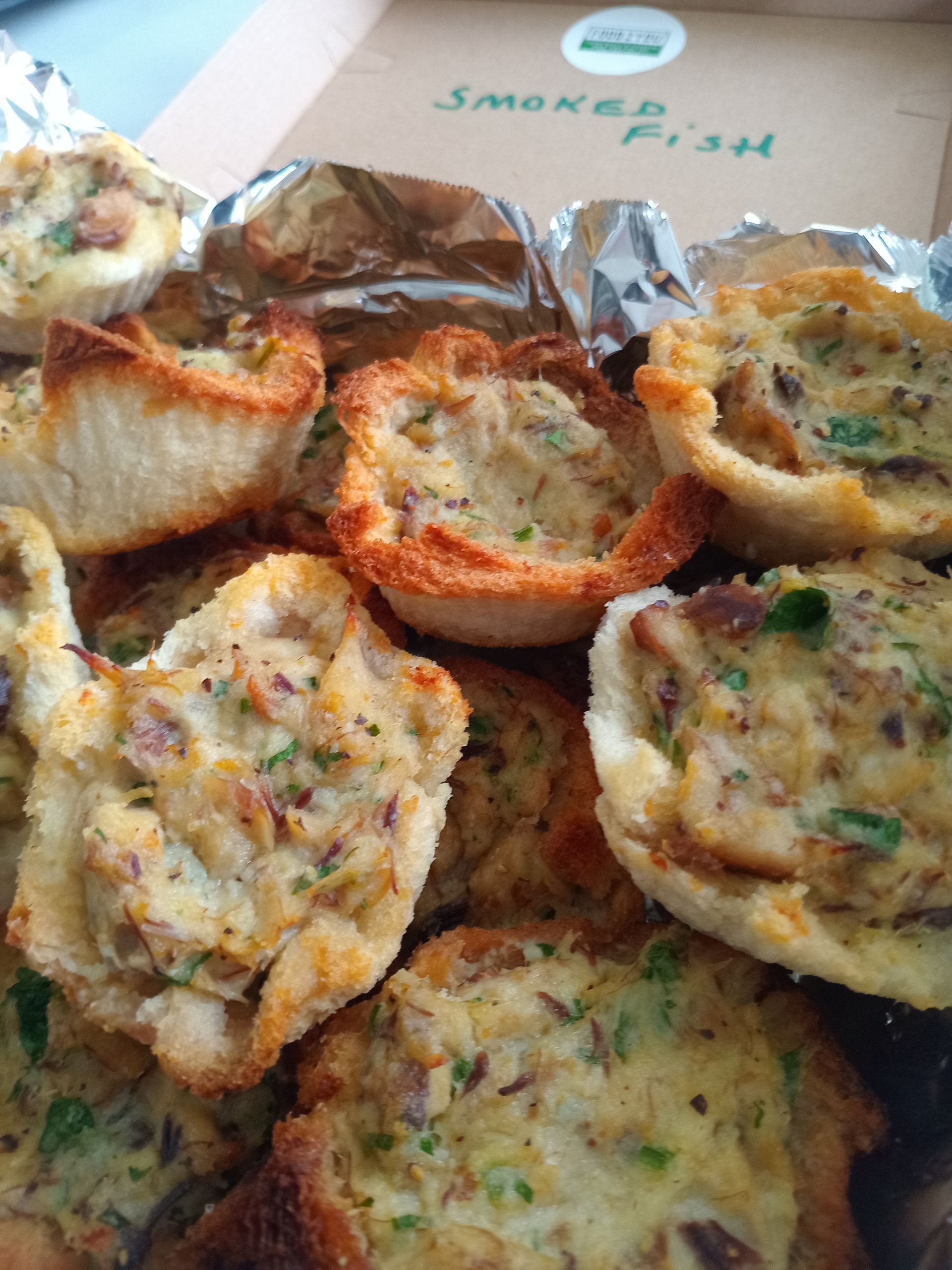 Close-up of many baked fish tarts in toasted bread cups, filling visible with parsley, in an open cardboard box.