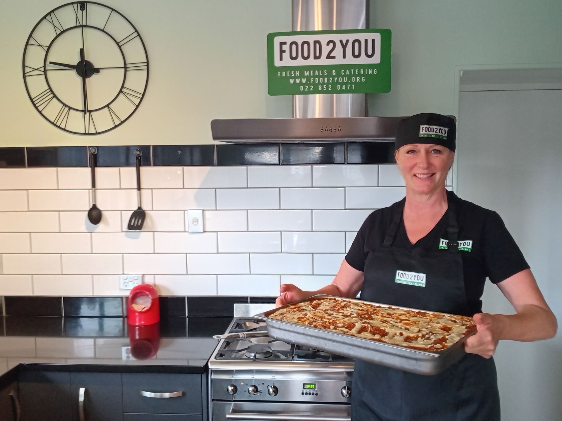 Woman in chef's hat and apron holding a baked lasagna in a kitchen, near a clock and FOOD2YOU sign.