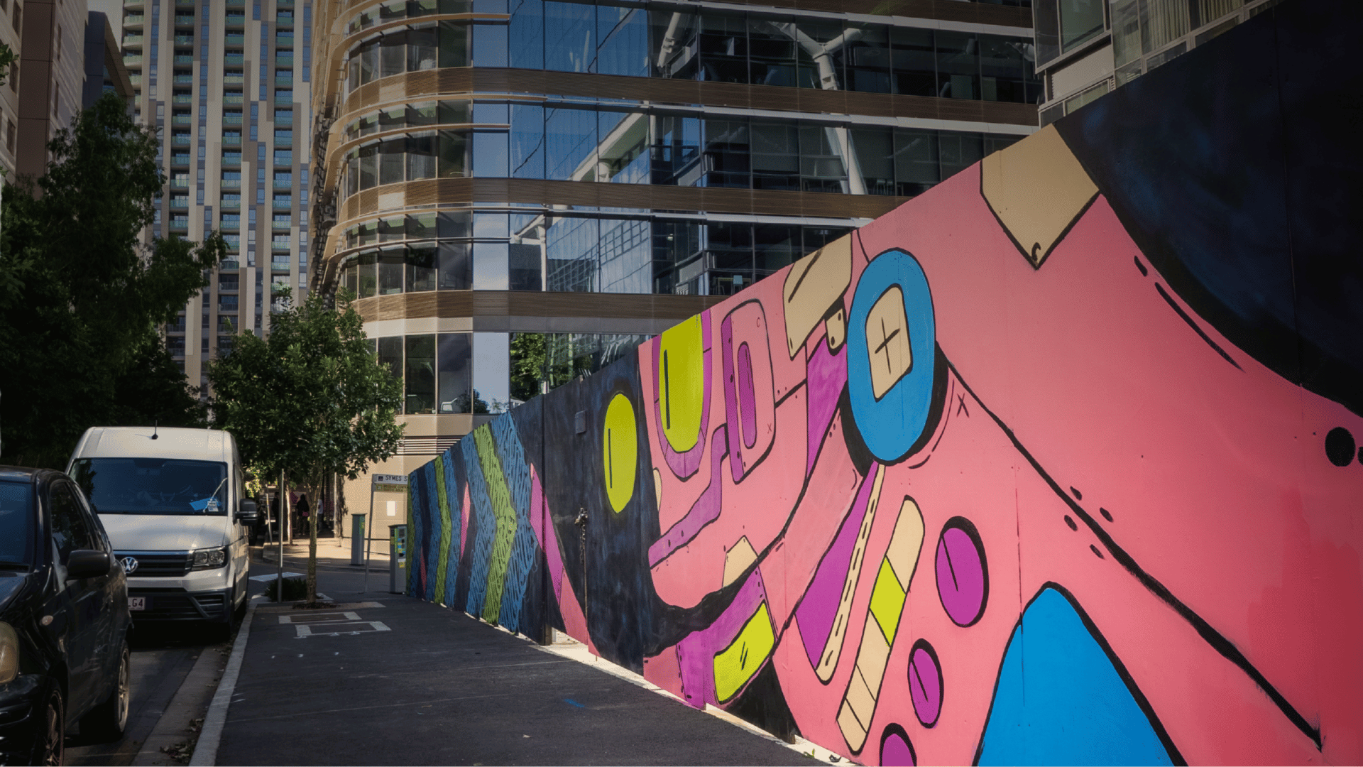 Colourful abstract mural on a wall next to a street with parked vehicles and a modern building in the background.