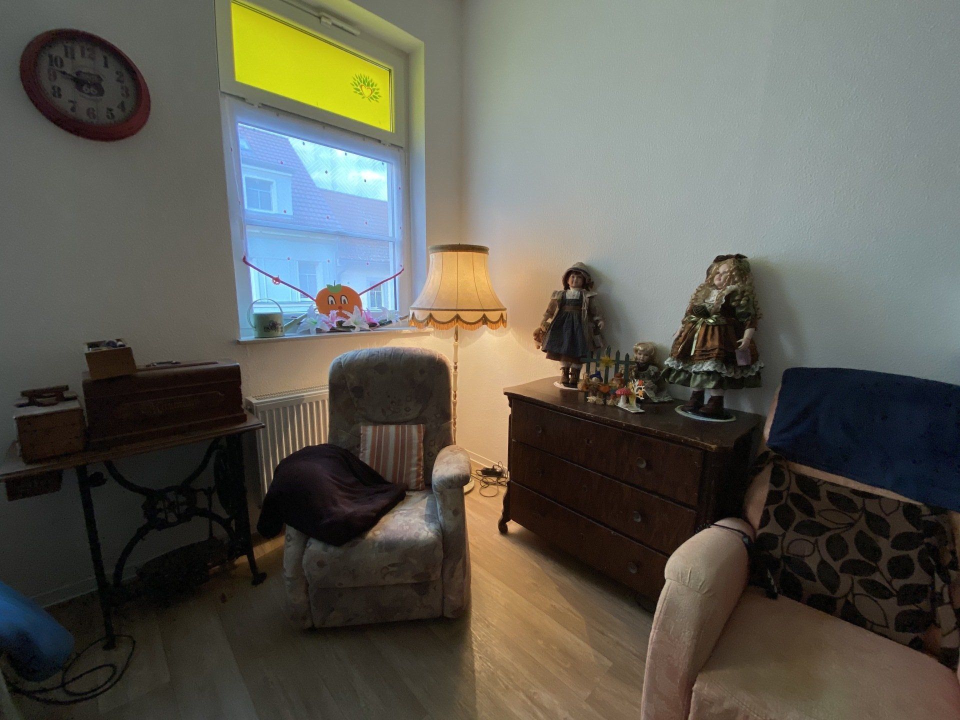 Gemütliches Zimmer mit antiken Möbeln, Puppen, einer Lampe und einem Fenster mit Tageslicht.