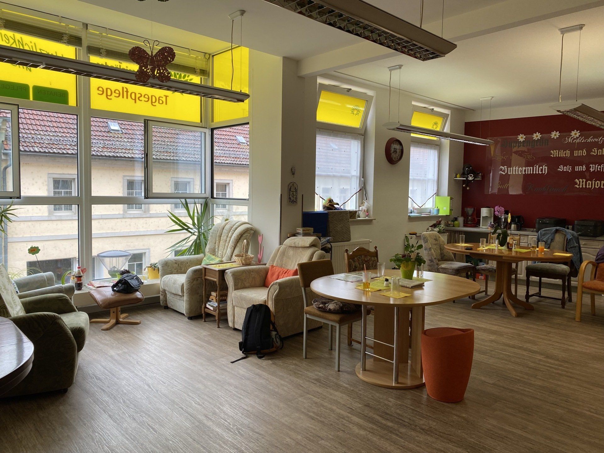 Ein gut beleuchteter Raum mit Sesseln, Tischen und großen Fenstern. Gelbe Akzente und rote Wanddekoration.
