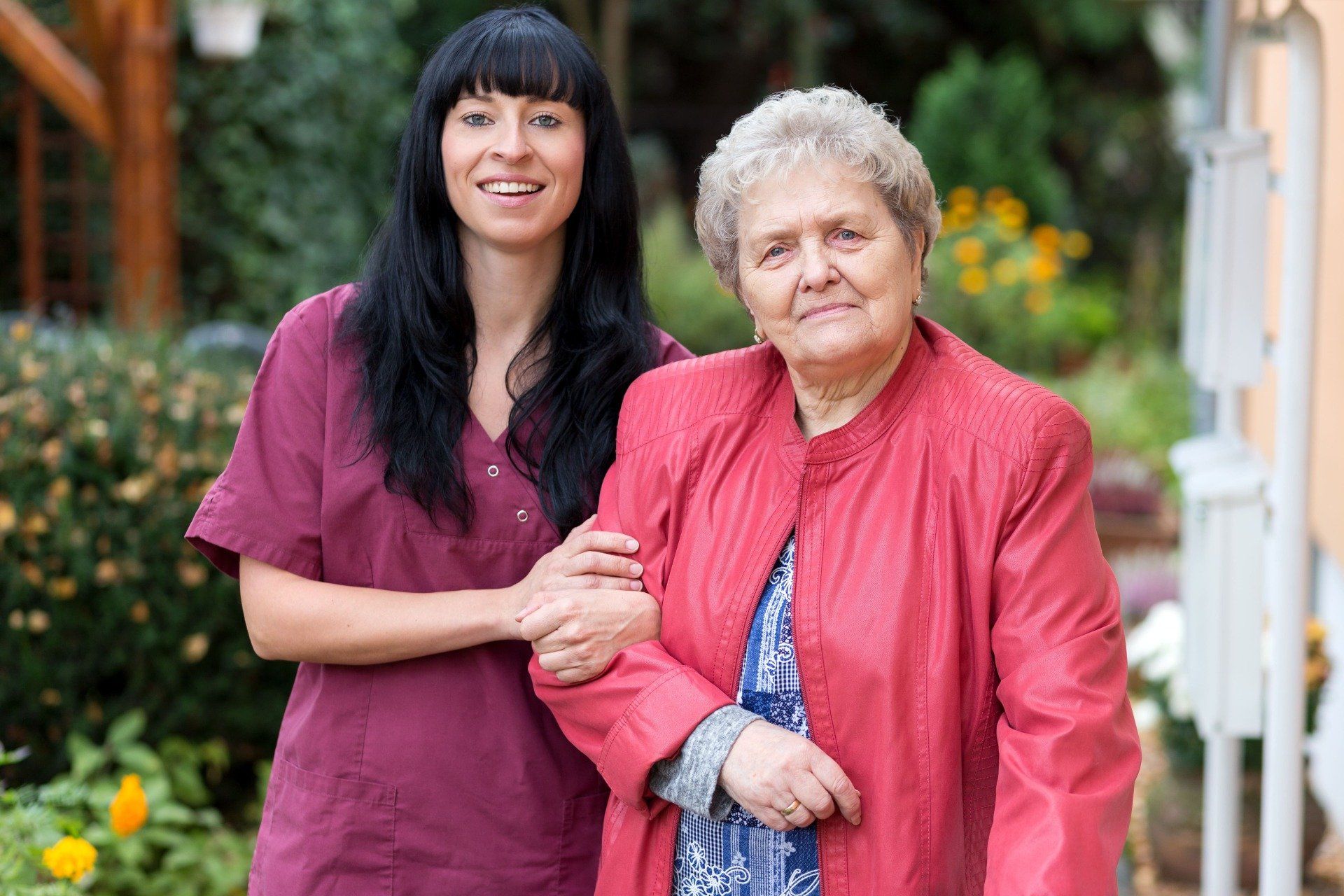 Frau im OP-Kittel hilft älterer Frau beim Gehen im Freien.
