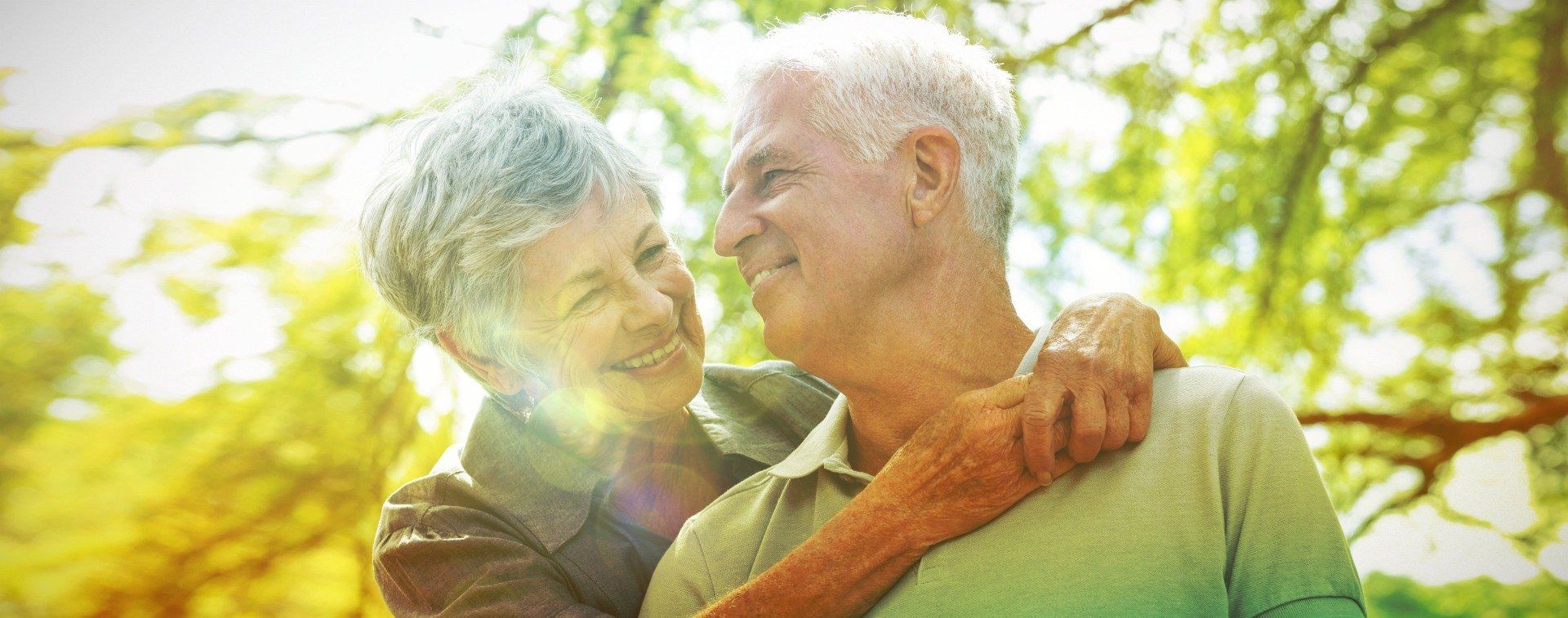 Älteres Paar umarmt sich im Freien und lächelt einander an. Umgeben von Grün und warmem Sonnenlicht.