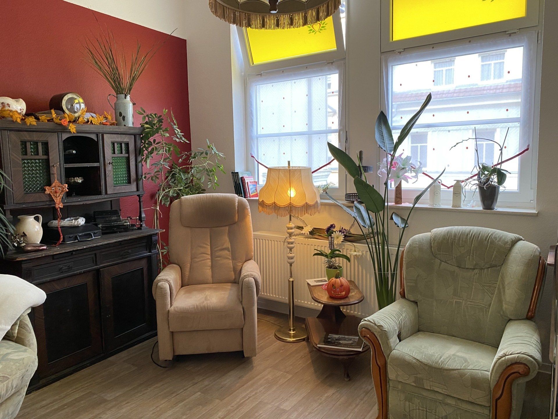 Gemütliches Wohnzimmer mit Sesseln, antikem Schrank, Lampe und Pflanzen in der Nähe eines Fensters mit gelben Akzenten.
