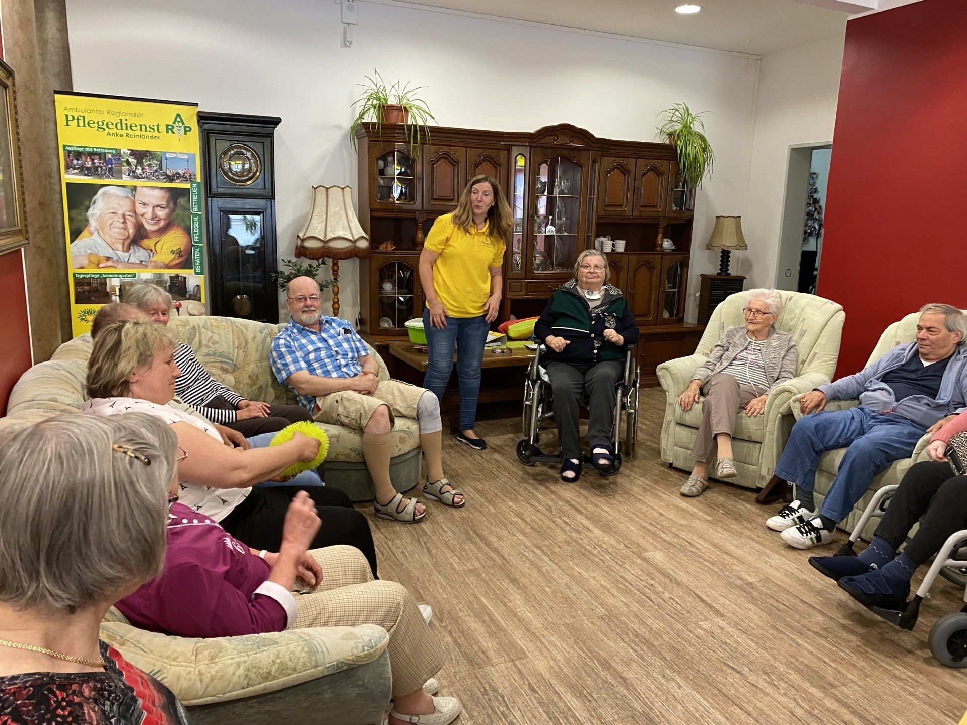 Eine Gruppe von Menschen sitzt und hört einer stehenden Person zu. Drinnen, heller Raum.