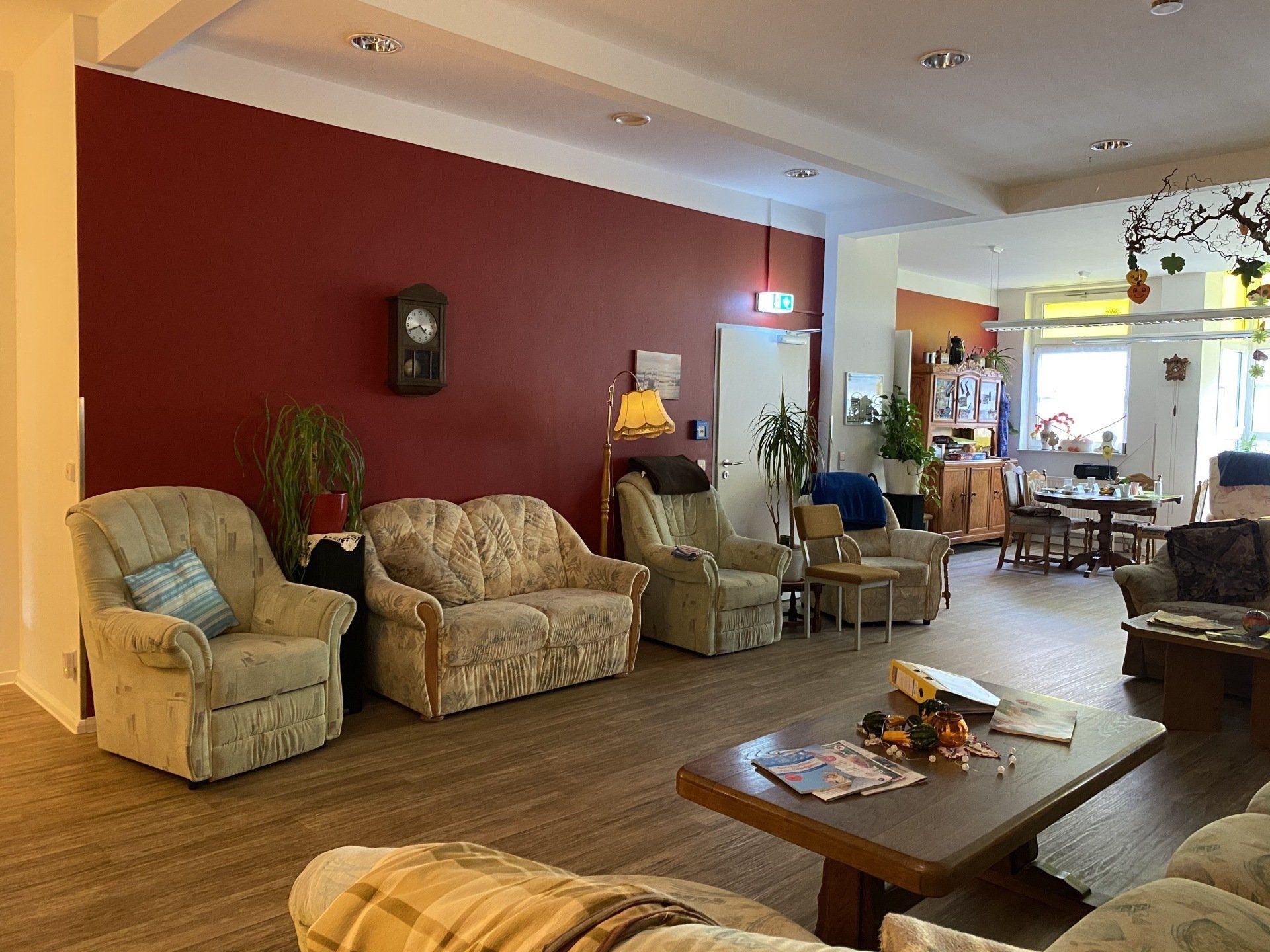 Wohnzimmer mit Sitzgelegenheiten, roter Akzentwand, Uhr, Pflanzen und Couchtisch.