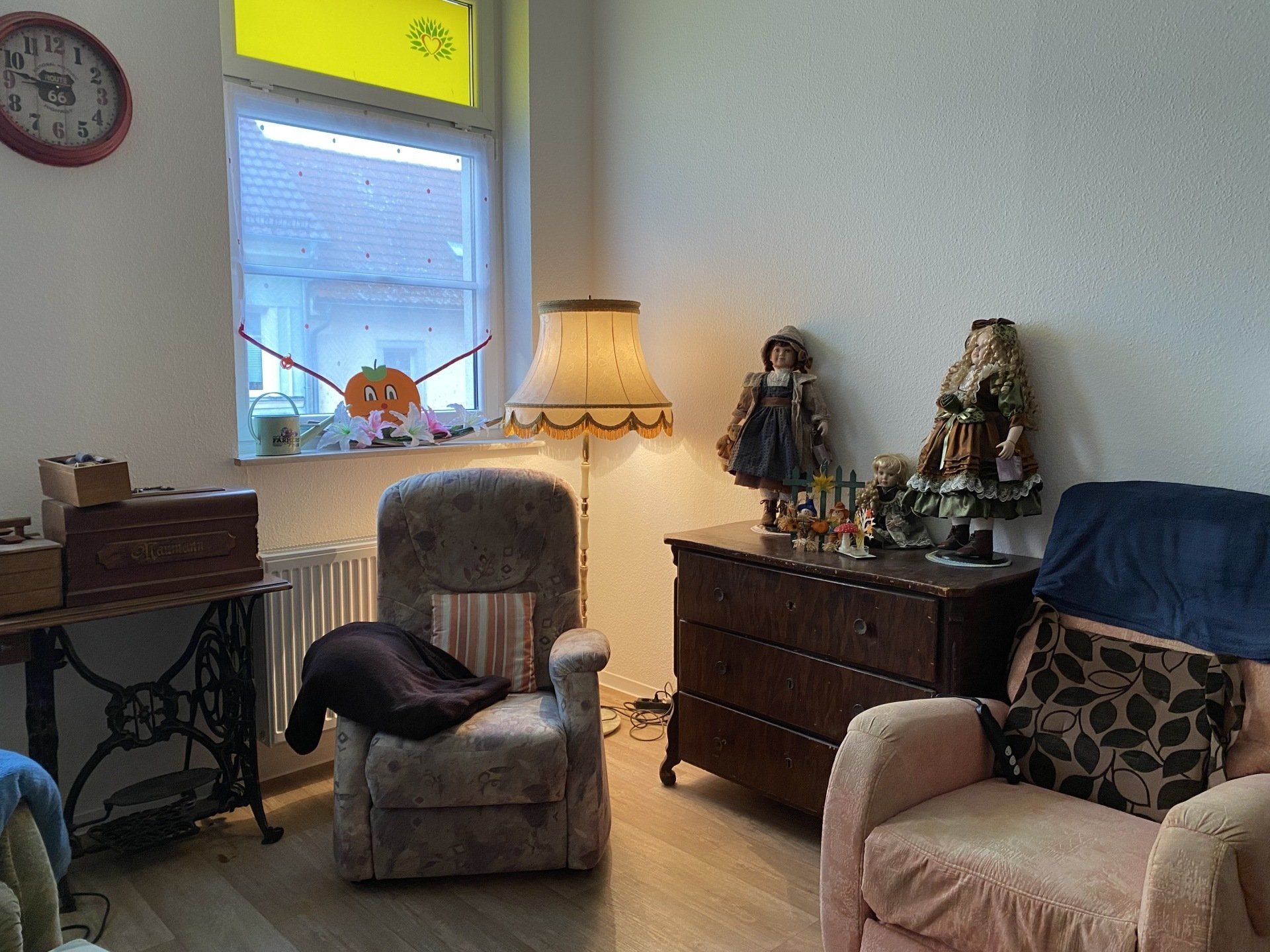Gemütliches Zimmer mit Sessel, Lampen, Kommode mit Puppen, Nähmaschinentisch und Fenster.