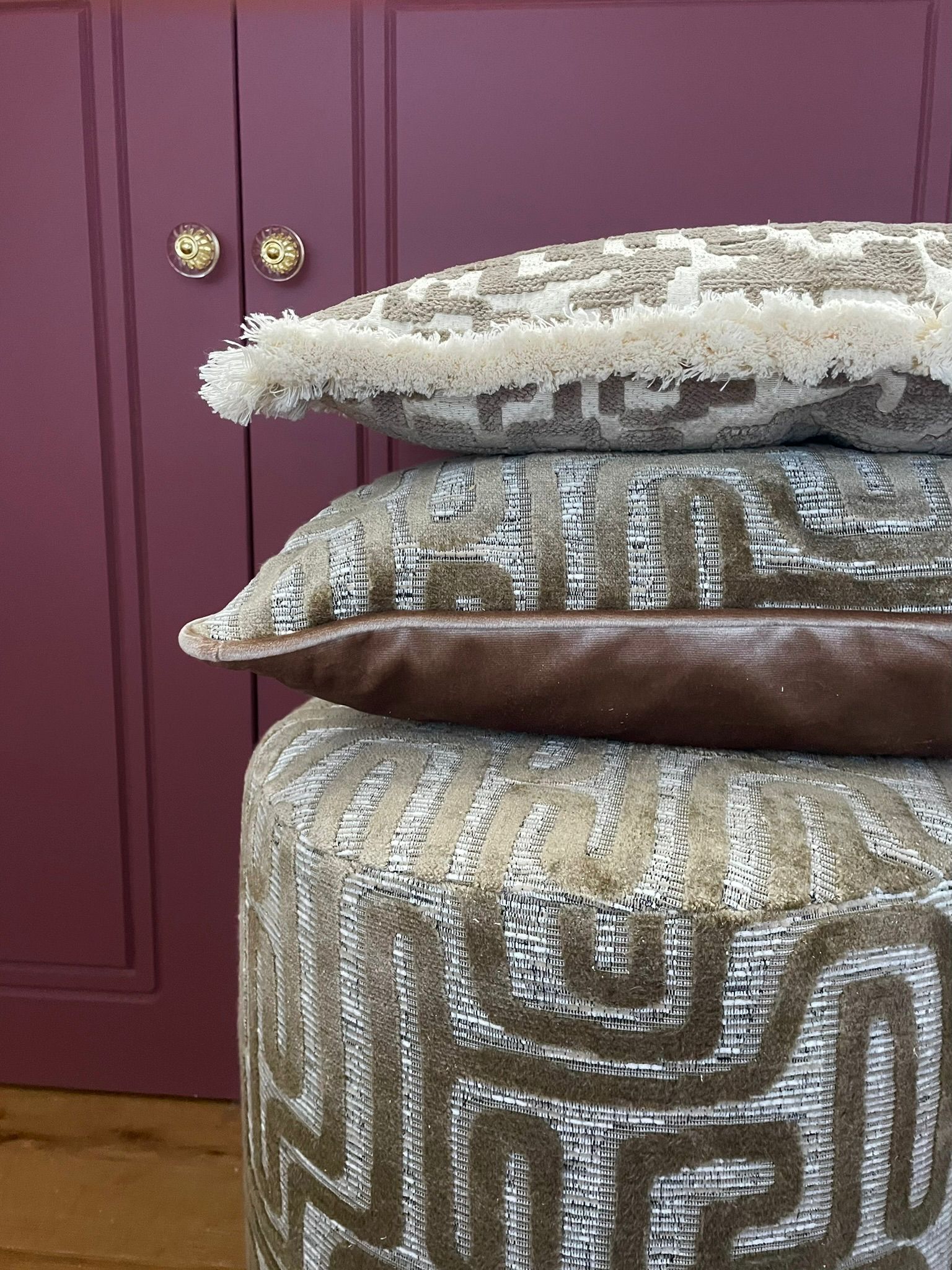 Een stapel sierkussens op een poef met patroon, voor een bordeauxrode kast.