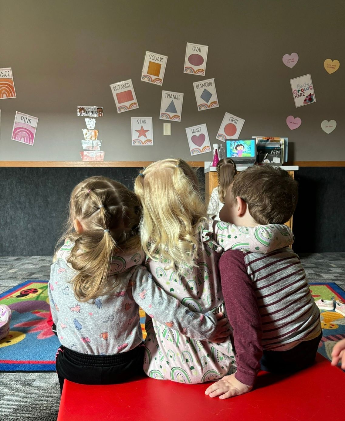 Three children are sitting on a red mat in front of a classroom.