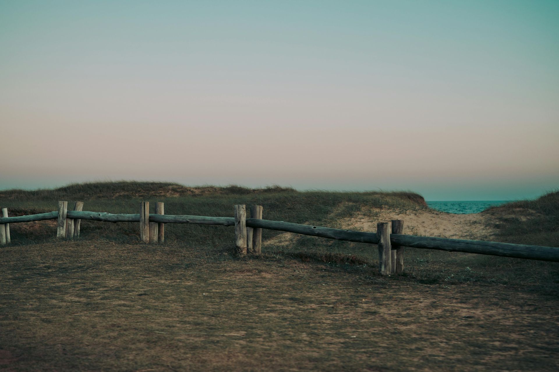 Wooden fence on grassy dune; ocean in distance; pastel sky.