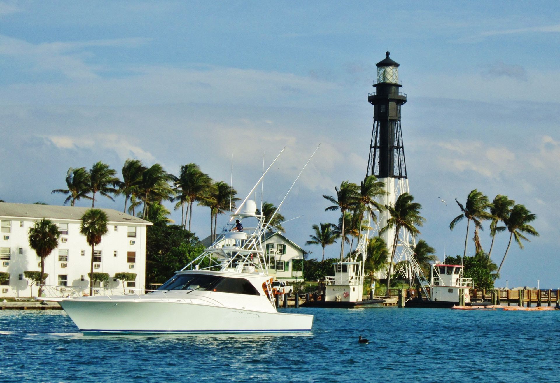 White yacht on blue water near a lighthouse and palm trees under a clear sky