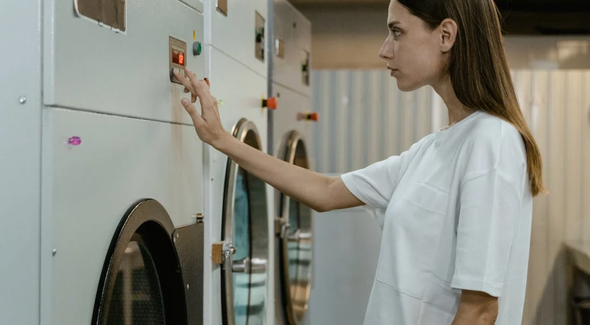 A woman is pressing a button on a washing machine in a laundromat.