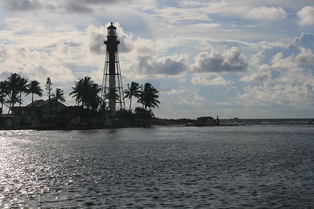 Coastal lighthouse on a palm-lined shore under cloudy sky, with calm water in the foreground