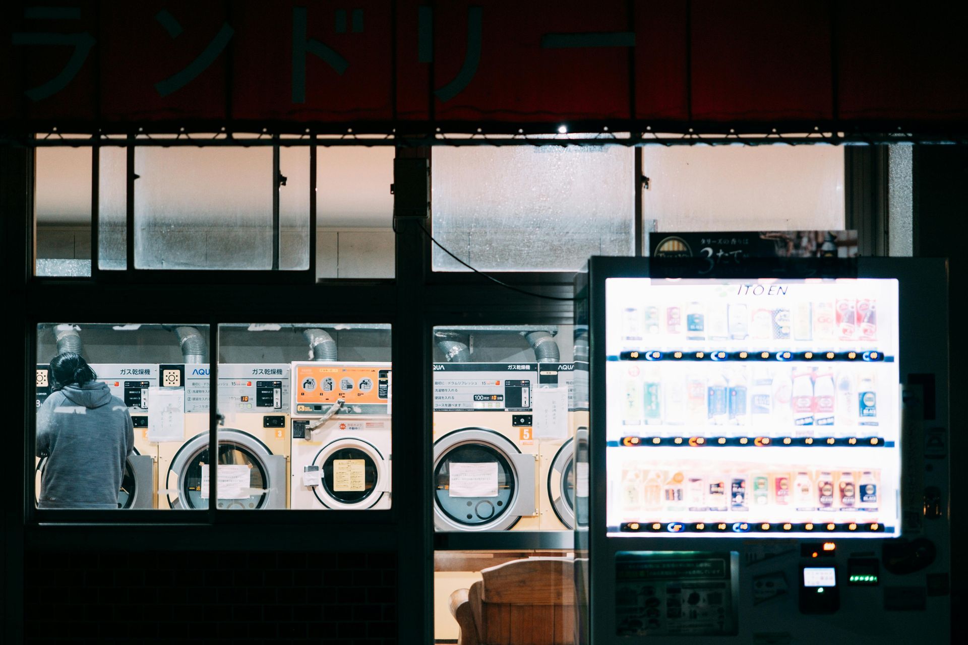Laundry storefront with lit vending machine and washing machines seen through dark windows