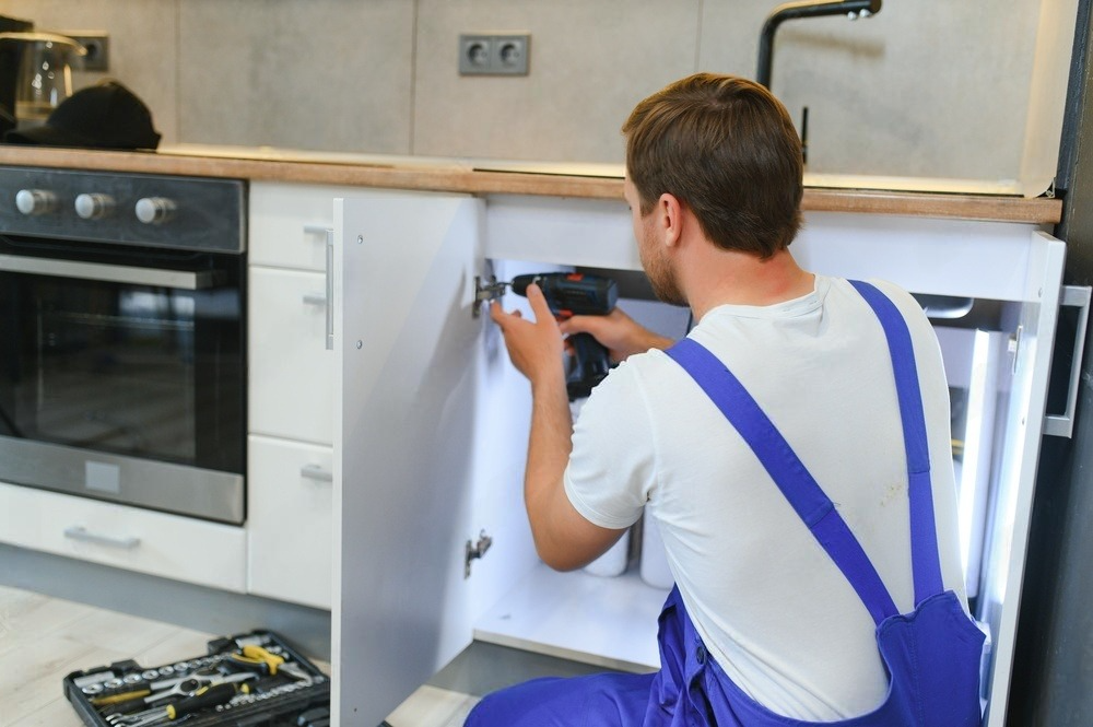 A man in blue overalls is fixing a sink in a kitchen.