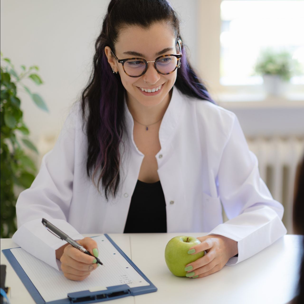 a woman in a lab coat is writing on a clipboard while holding an apple .