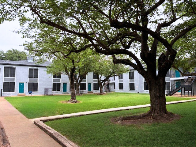 Apartment complex with white buildings, teal doors, and green lawn under large trees.