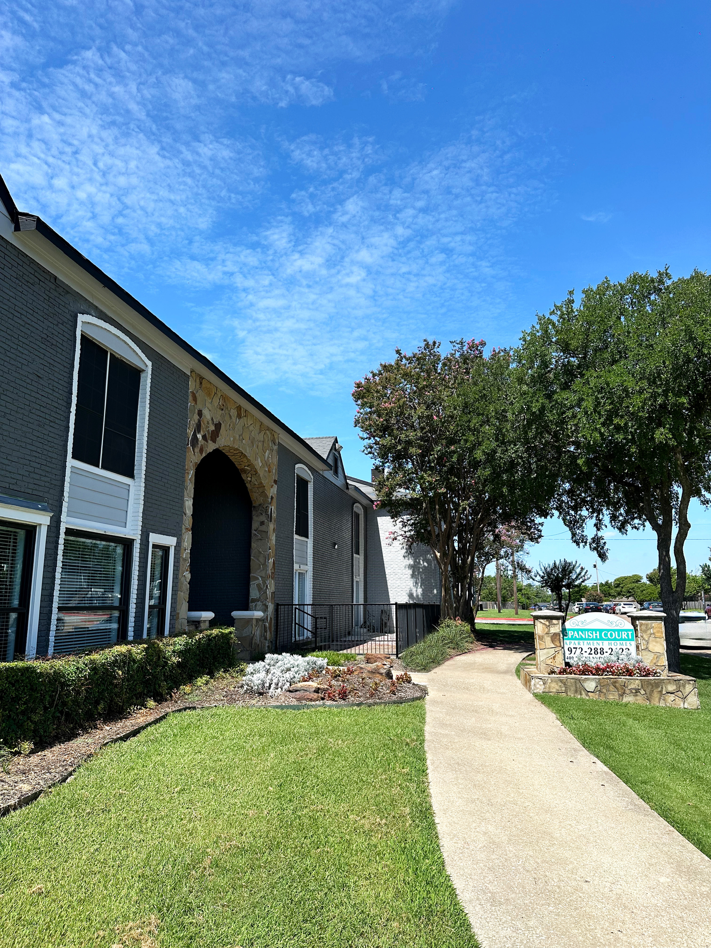 Exterior view of a building with gray siding and a walkway, lush green lawn, and blue sky.
