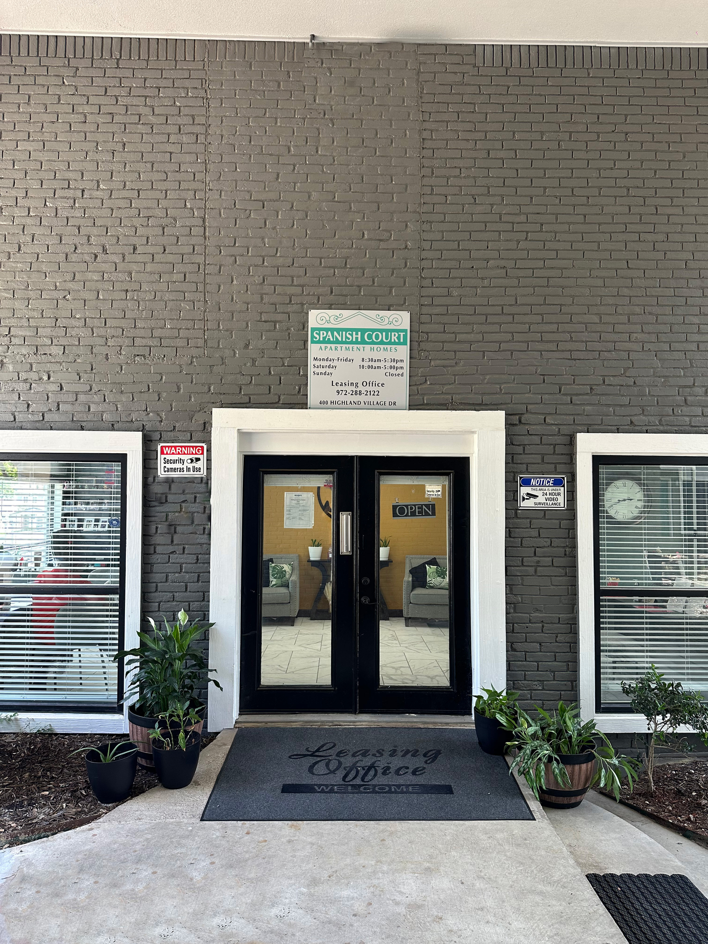 Gray brick building entrance with double doors, welcome mat, and small plants.