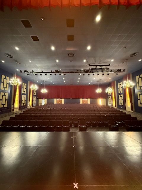 An empty auditorium viewed from the stage, featuring rows of dark seats, chandeliers, and a deep red stage curtain.