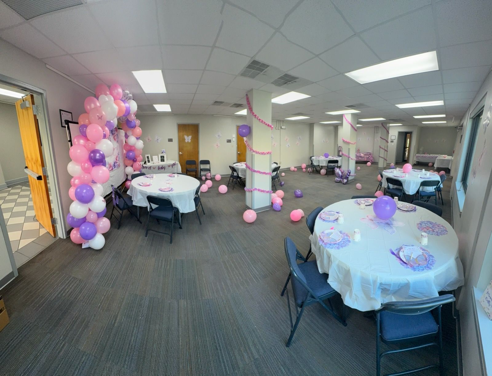 Party room with pink and purple balloons, round tables with white tablecloths, and folding chairs.