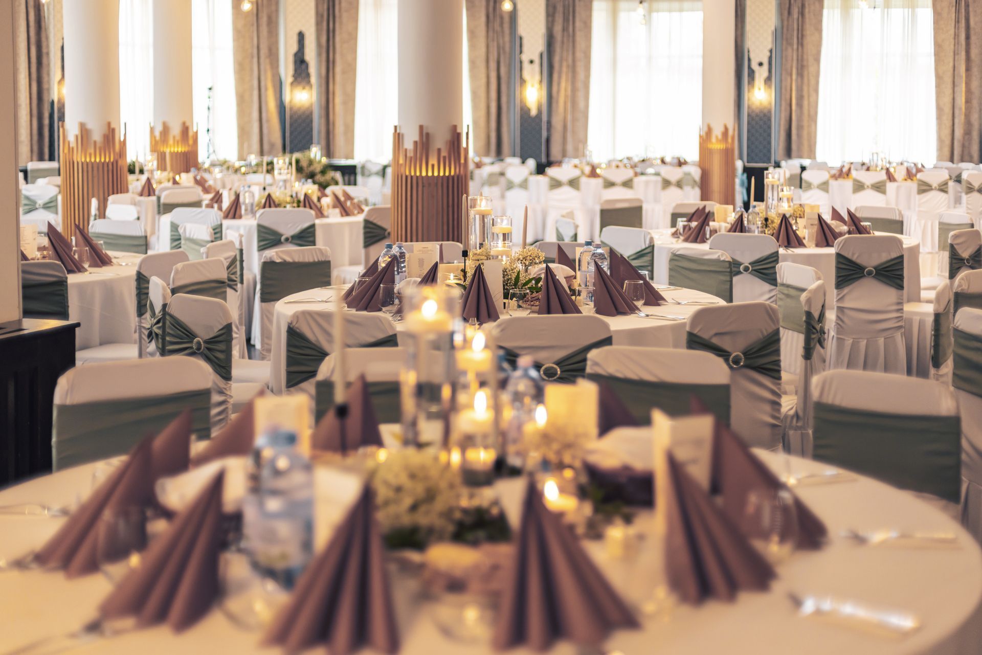 Banquet hall set with round tables, white tablecloths, olive green chair sashes, and mauve napkins.