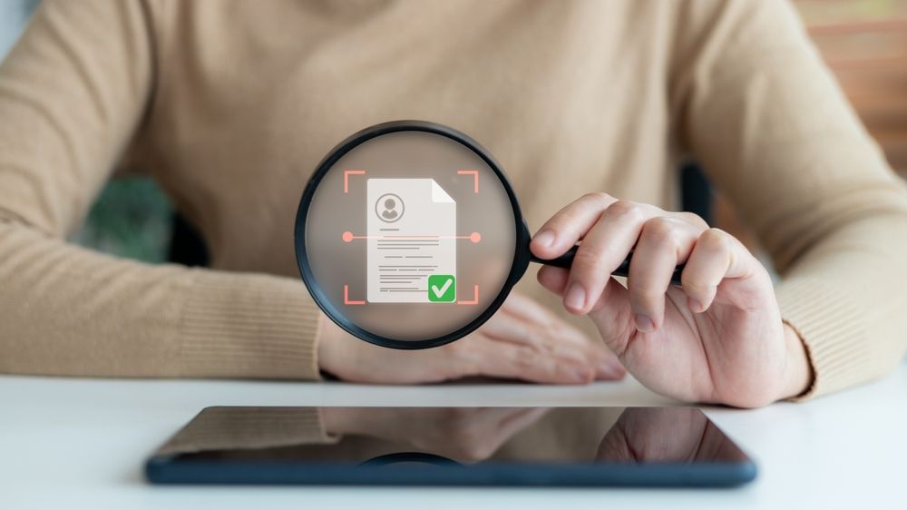 A person holding a magnifying glass over a digital document with a checkmark, representing document verification.