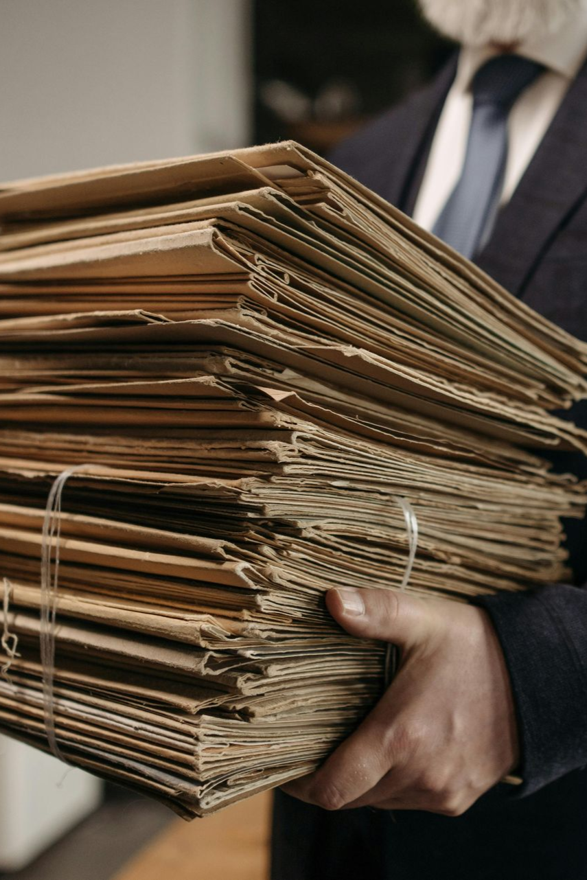 A person in a business suit holds a large, bundled stack of folded cardboard sheets.