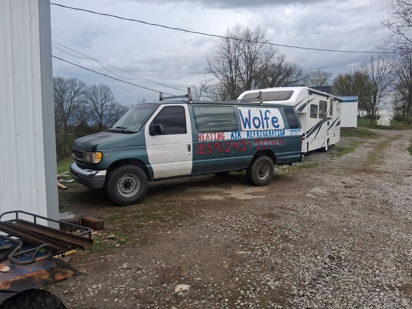 Blue-and-white service van with “Wolfe” sign parked on a gravel lot beside a white building under cloudy skies