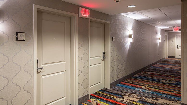 Hotel hallway with doors, carpeted floor, emergency exit sign, and wall sconces.