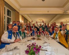 Eine jubelnde Gruppe an einer langen Festtafel winkt und lächelt in einem warm beleuchteten Speisesaal.