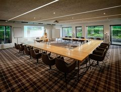Leerer Konferenzraum mit U-förmigem Holztisch, schwarzen Stühlen, Projektionsleinwand und großen Fenstern