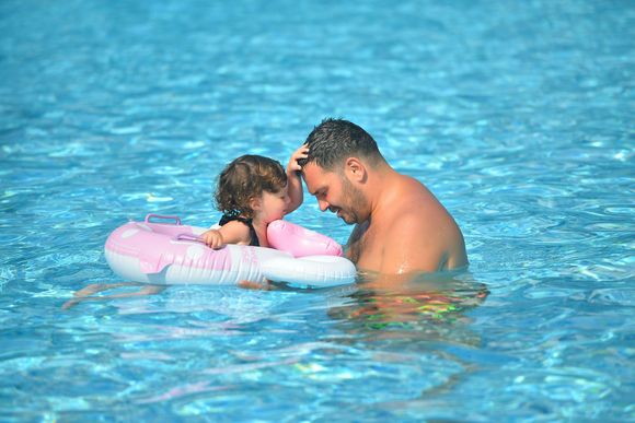 An adult and a child in an inflatable ring swim in a bright blue pool, with the child touching the adult’s head.
