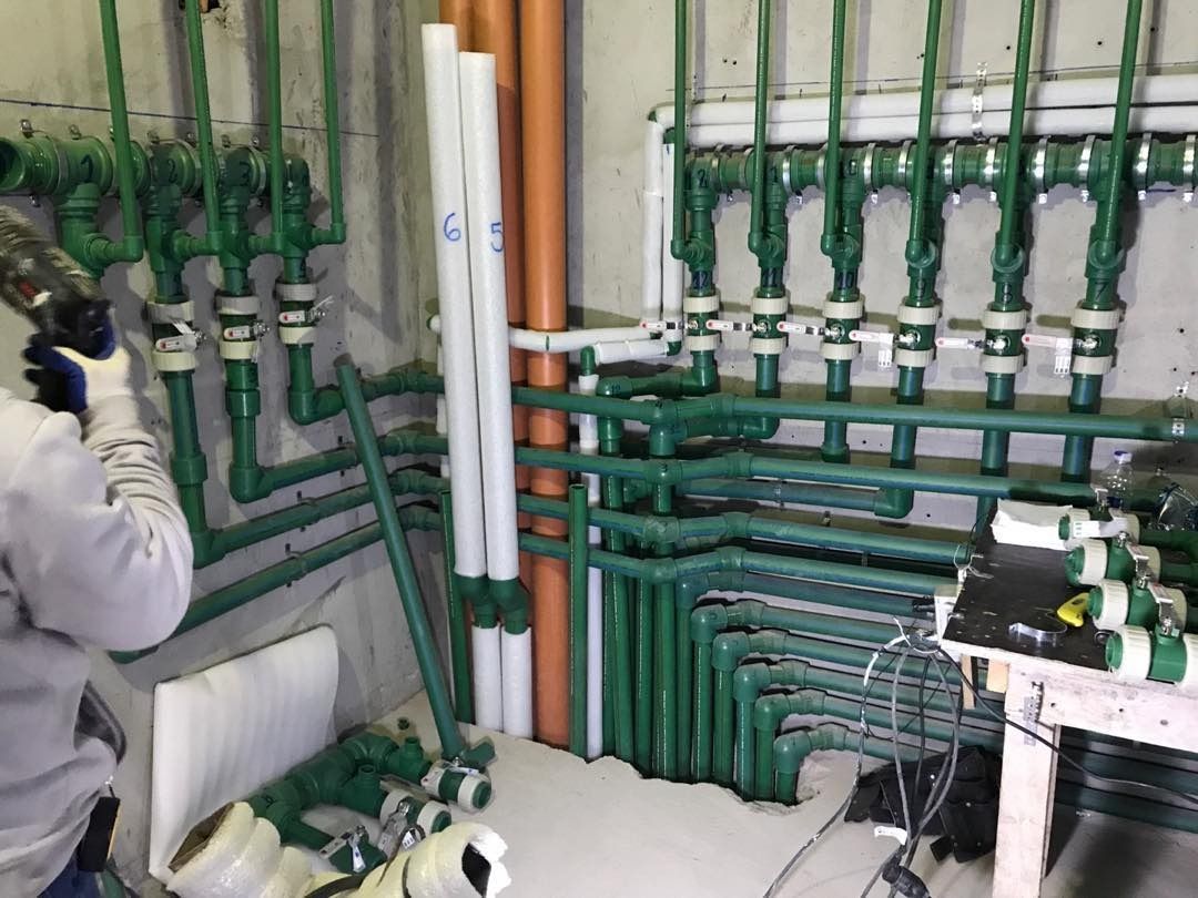 Tuberías de plomería en una habitación. Una persona con guantes usa una herramienta para limpiar las tuberías. Se ven tuberías verdes y naranjas.