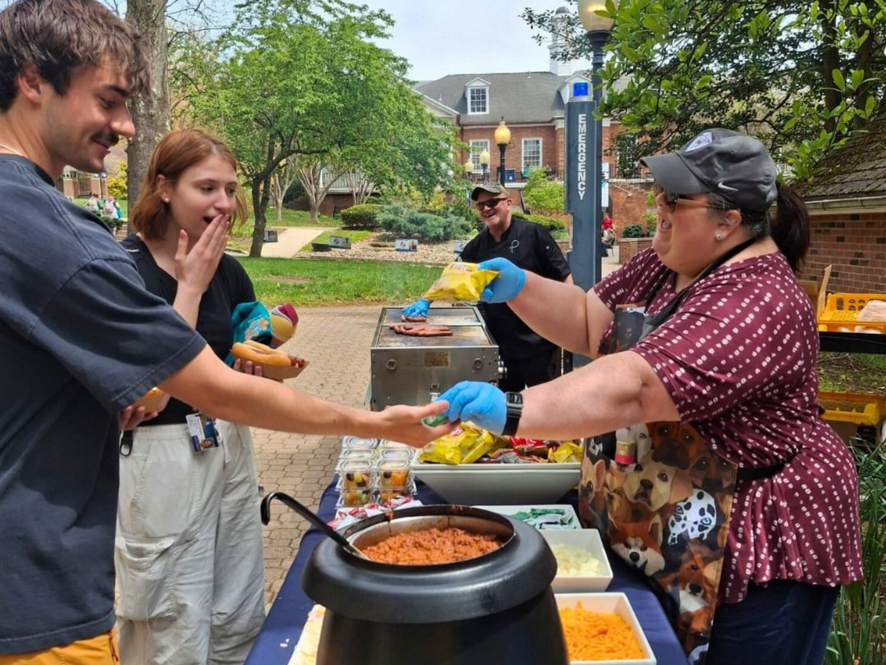 People receiving food at an outdoor event: chili, hot dogs, and toppings. Person smiling hands out snacks.