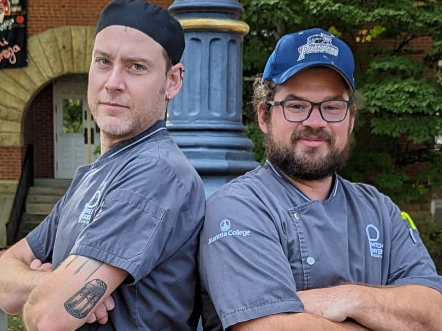 Two chefs stand back to back, arms crossed. One wears a black cap, the other a blue baseball cap.