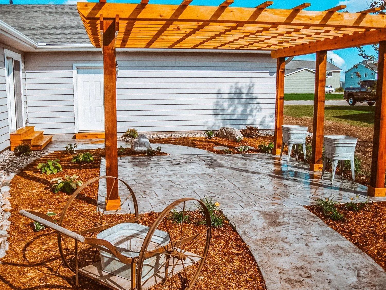 Wooden pergola over a stamped concrete patio, next to a building. Two white chairs and a garden path are in view.