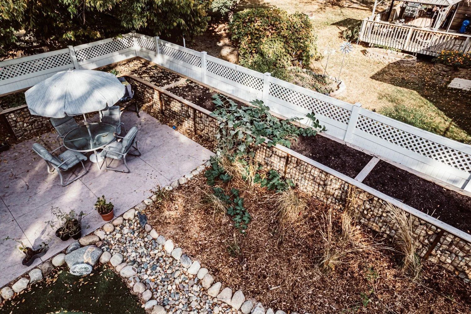 Patio with a table and chairs under an umbrella, surrounded by a garden and a white lattice fence.