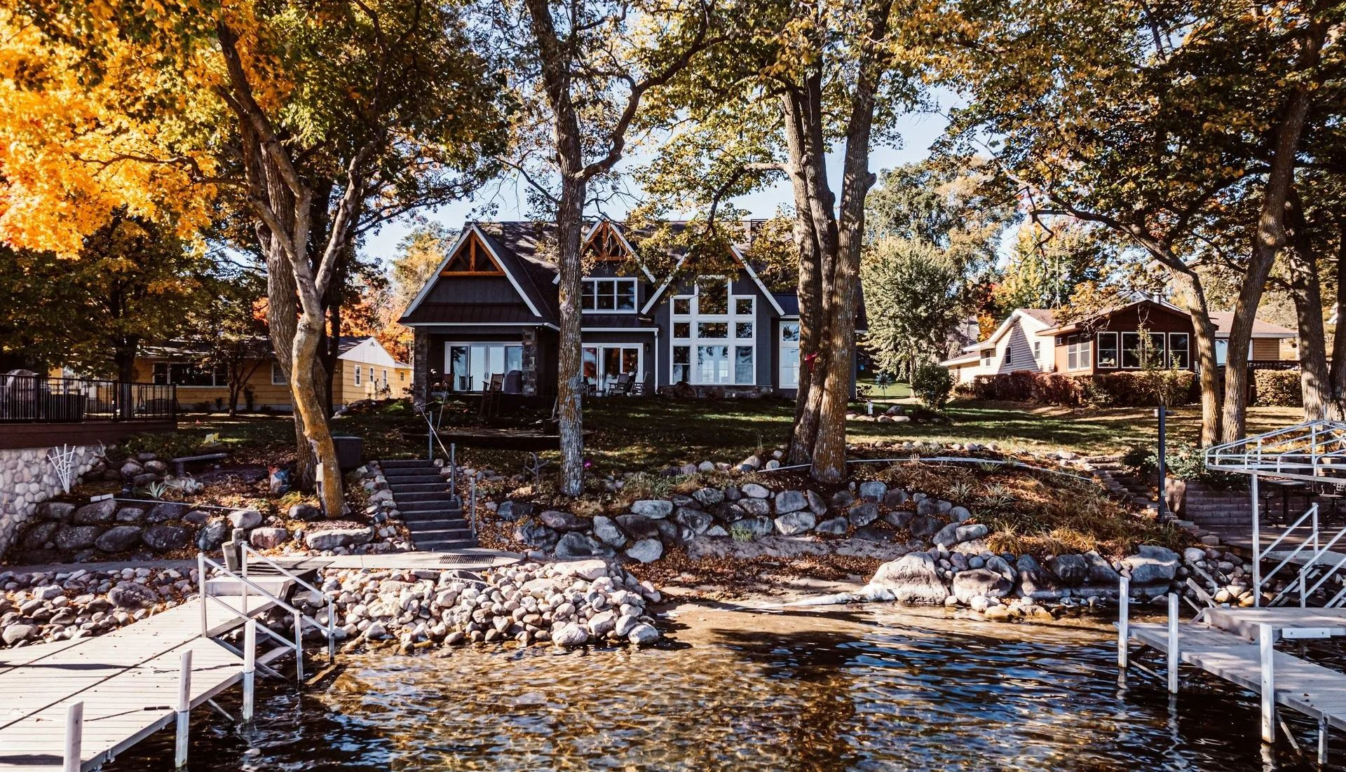 House on a lake shore surrounded by trees. Steps lead up to the dark-colored house from the dock.