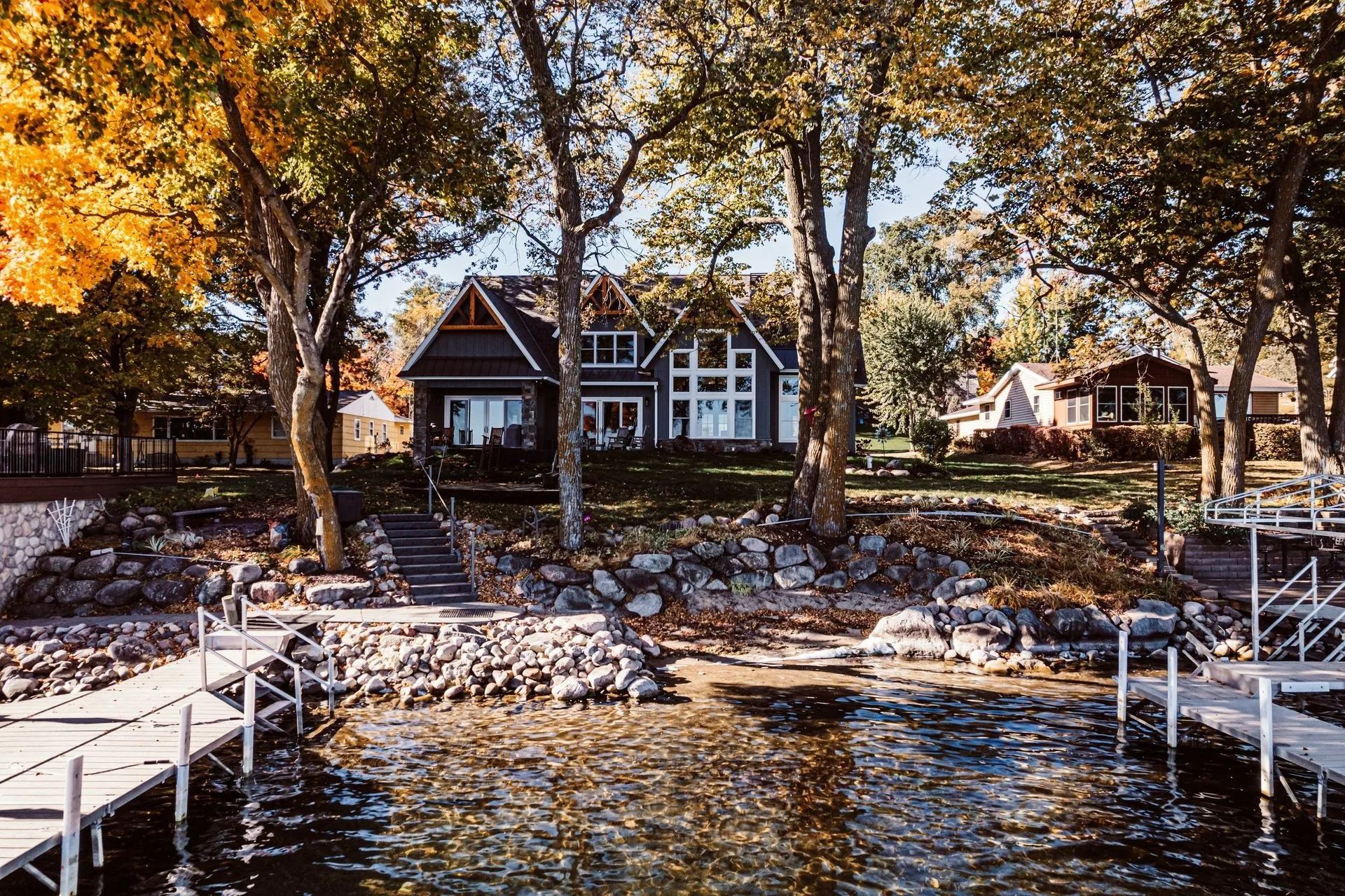 Lakefront home with large windows, surrounded by trees with autumn foliage, and two docks.