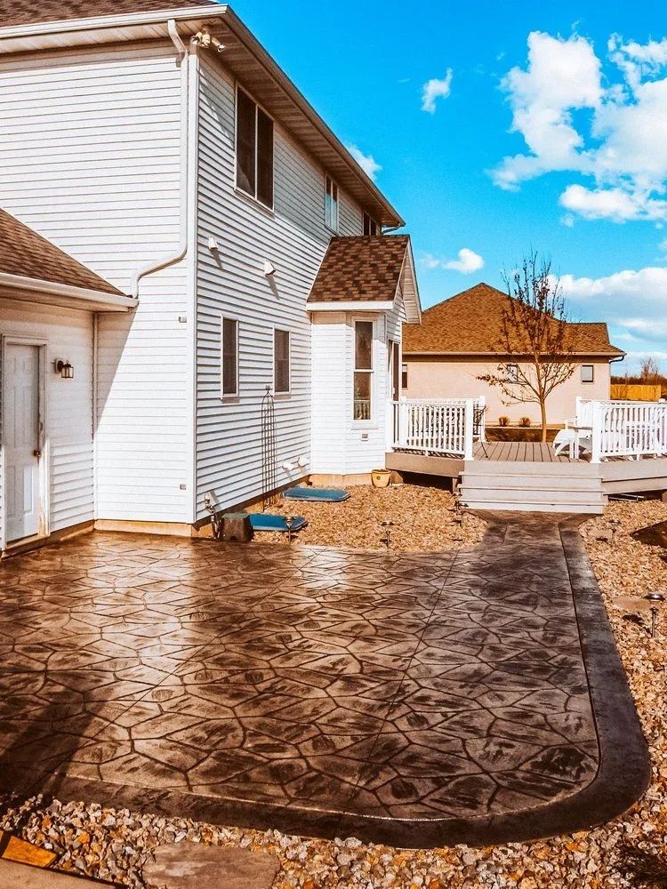 Backyard patio with stamped concrete, gravel border, and two-story white house with brown roof against blue sky.