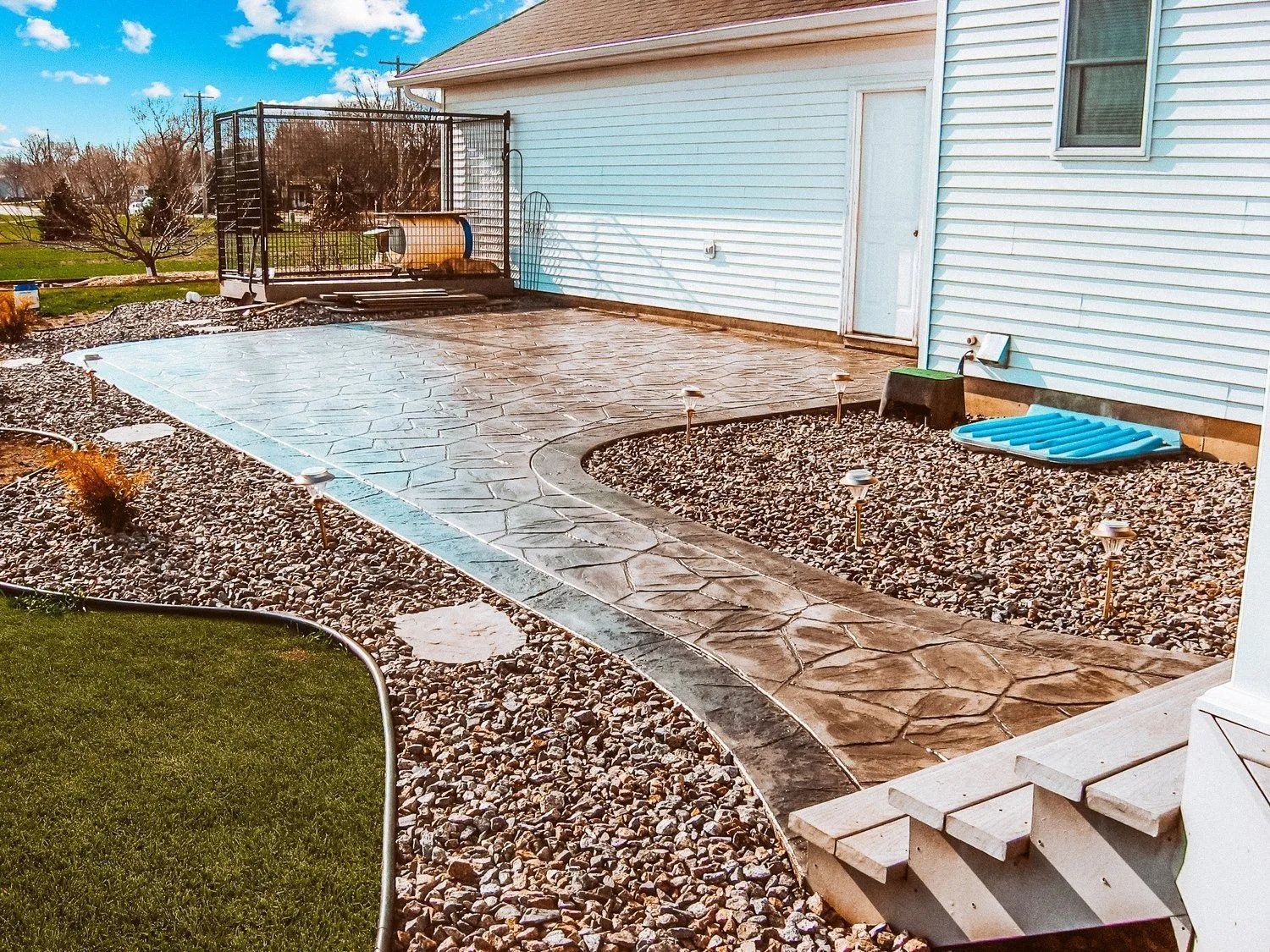 Stamped concrete patio with a curved path, surrounded by gravel, next to a white house with a lawn.