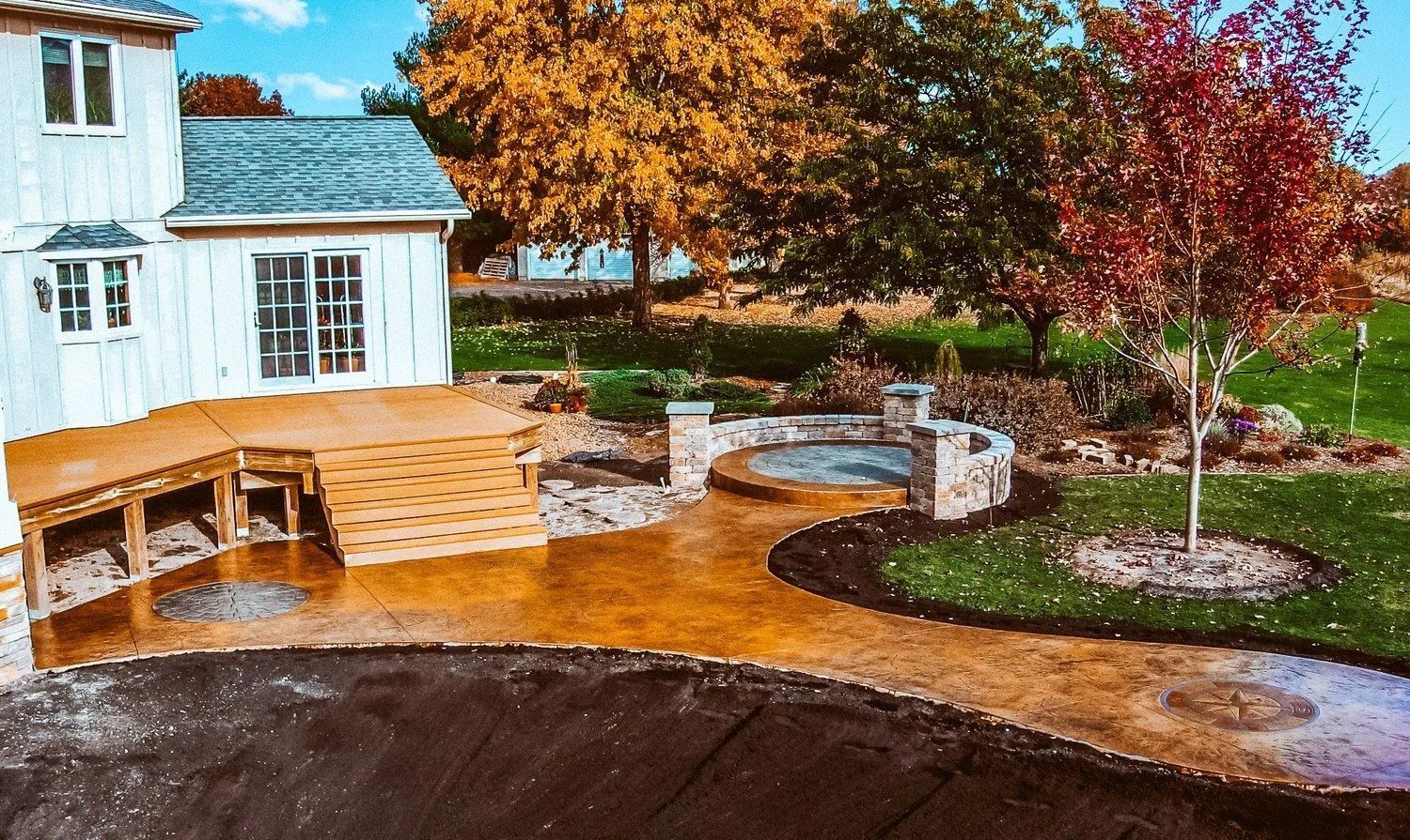 Backyard with a deck, concrete path, and circular stone patio near a two-story white house and colorful trees.
