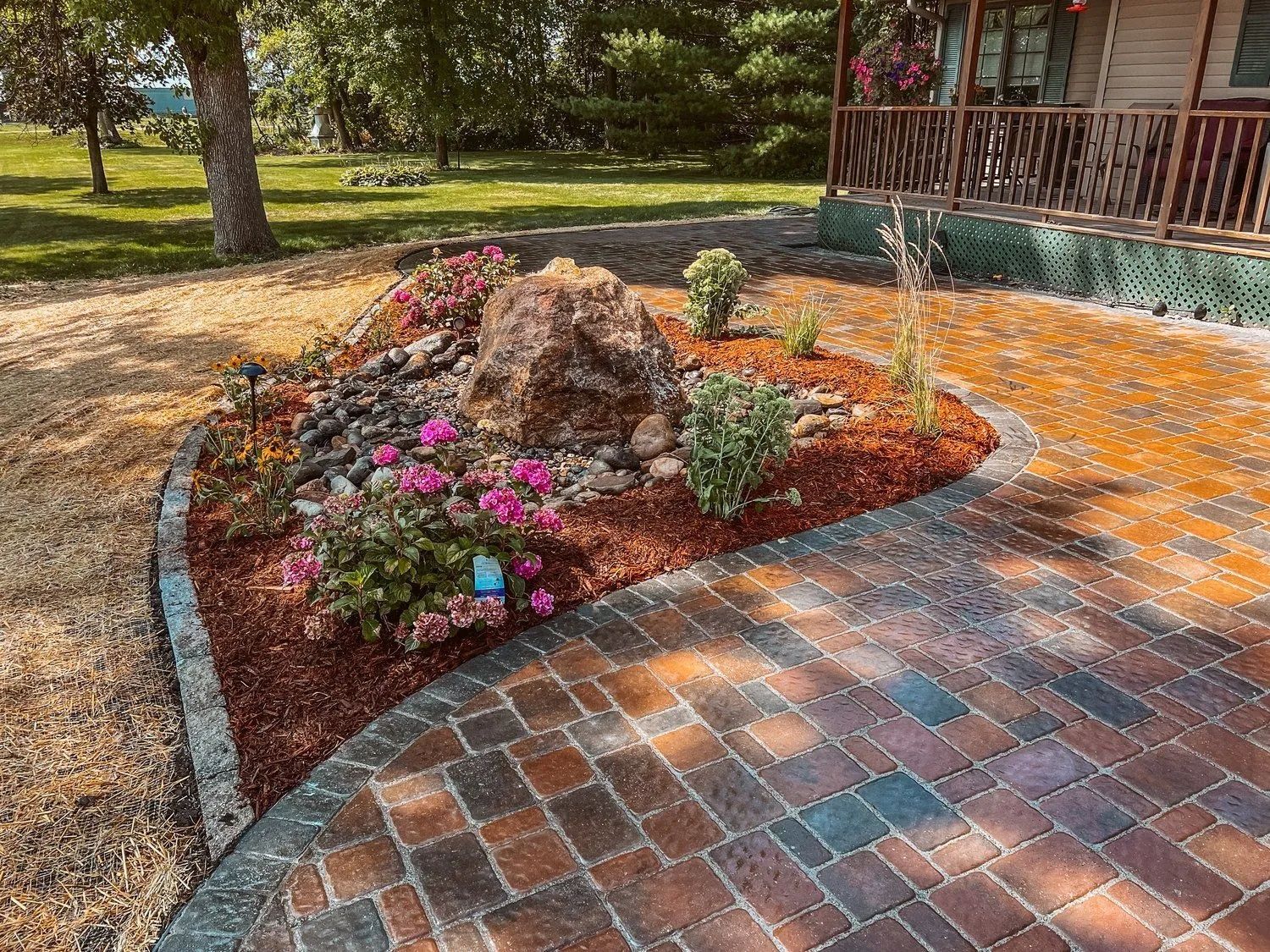 Landscaped garden bed with pink flowers, large rock, and brick pathway near a house.