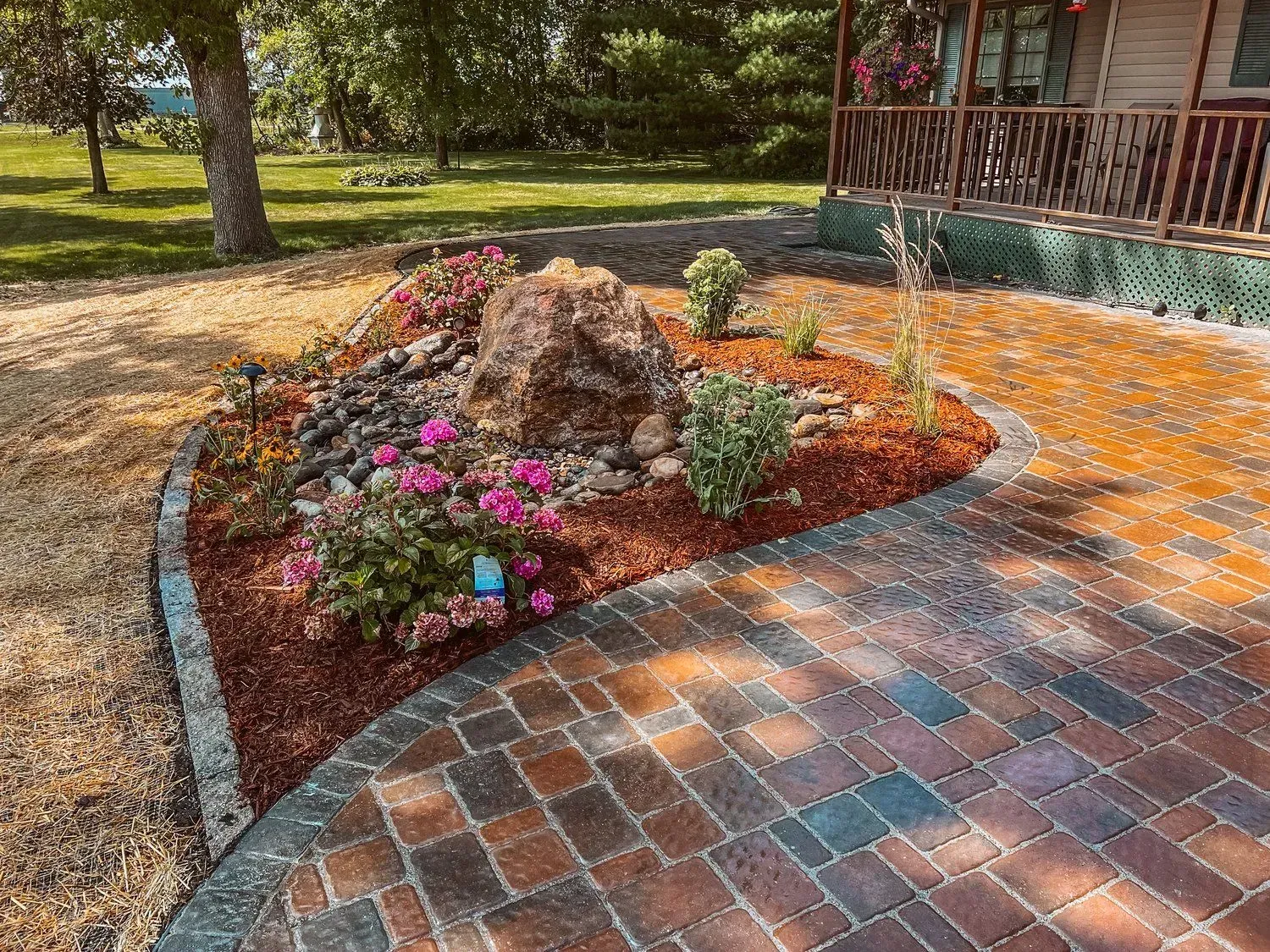 Brick walkway curving around a landscaped garden bed with large rock, flowers, and a house in the background.
