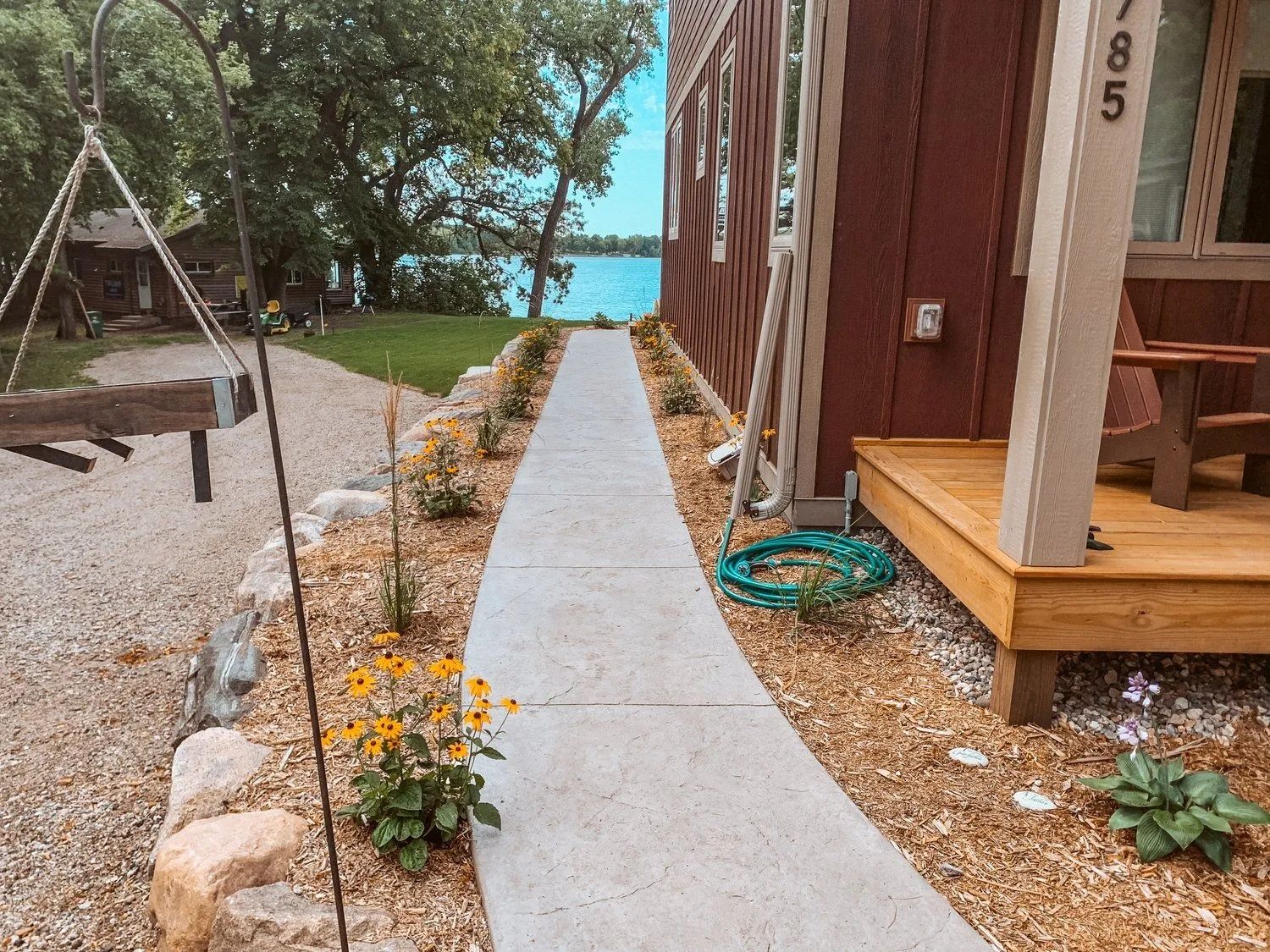 Concrete walkway lined with plants leads to a lake. A red-brown building and gravel path are visible.